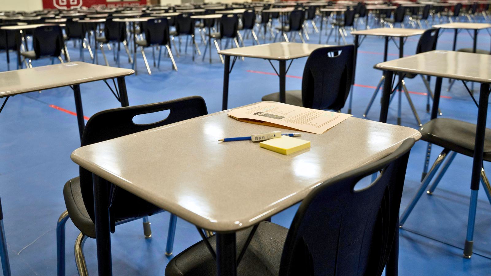 Empty exam desks in a school gym with a test paper and pencil on one desk.